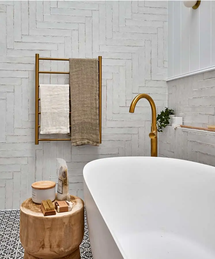 Modern bathroom with white tub, brass fixtures, wooden stool, and textured white walls with towels on gold rack.
