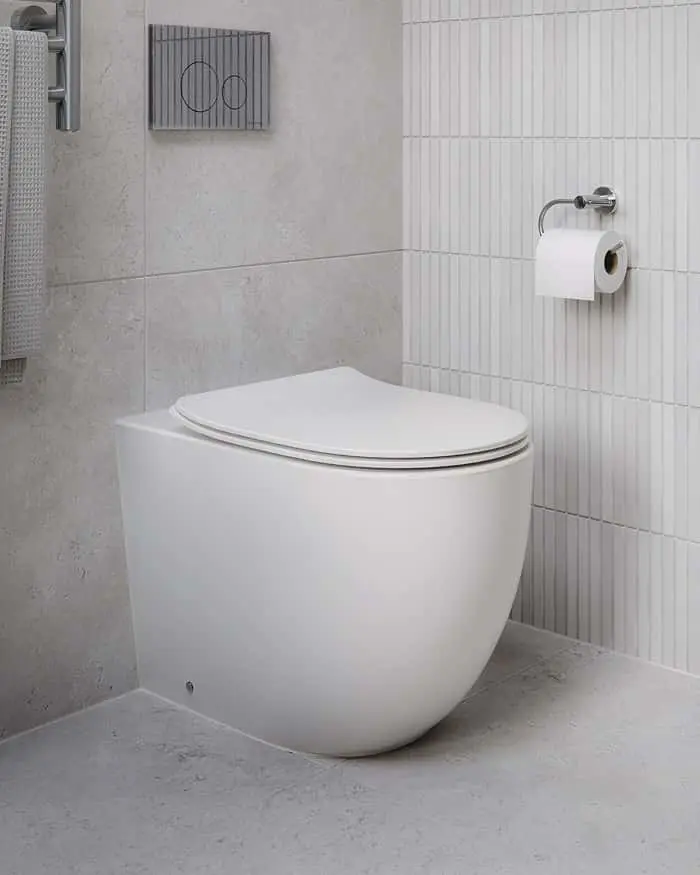 Modern white toilet in bathroom with gray and white textured tiles, toilet paper holder, and flush plate on wall.