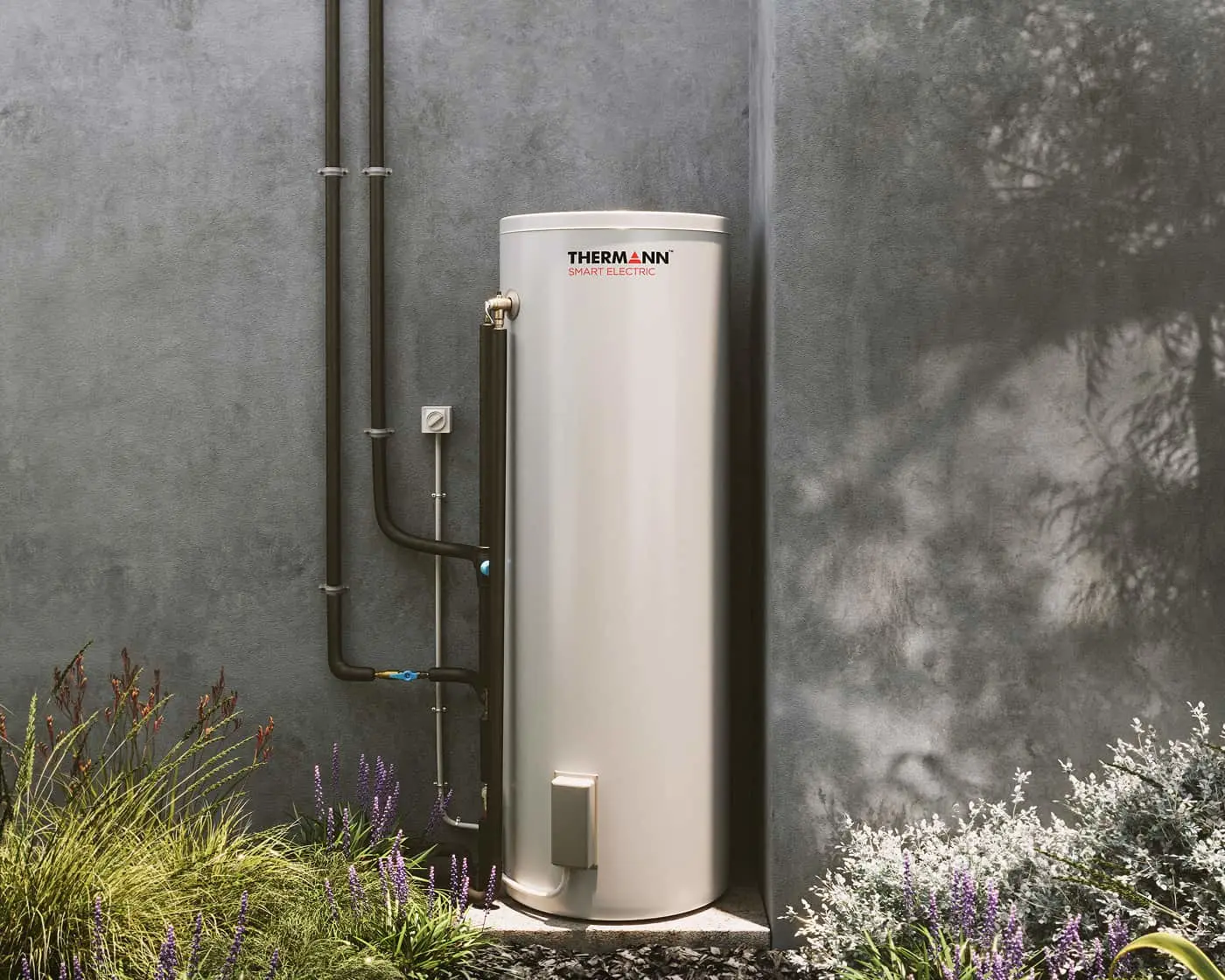 White Thermann Smart Electric water heater mounted on gray concrete wall, surrounded by purple and green garden plants.