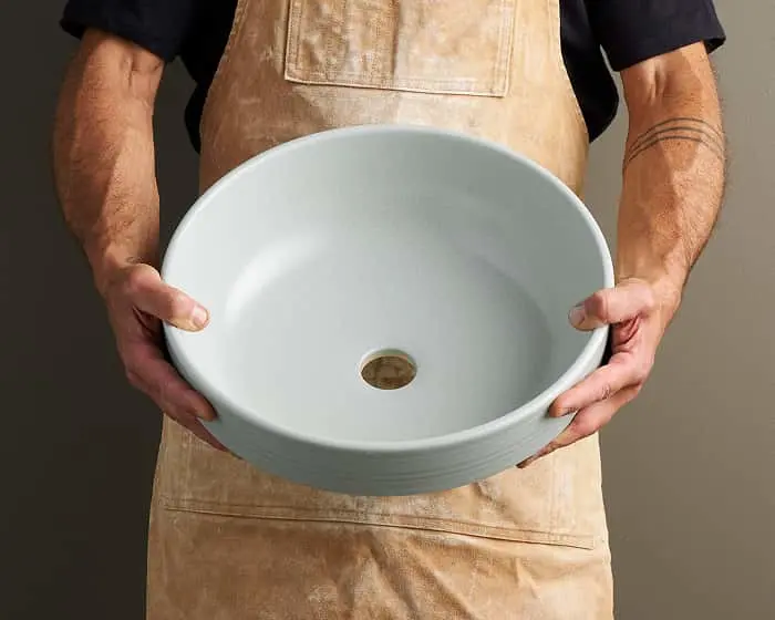 Person in tan apron holding a round white ceramic sink basin with drain hole in center.