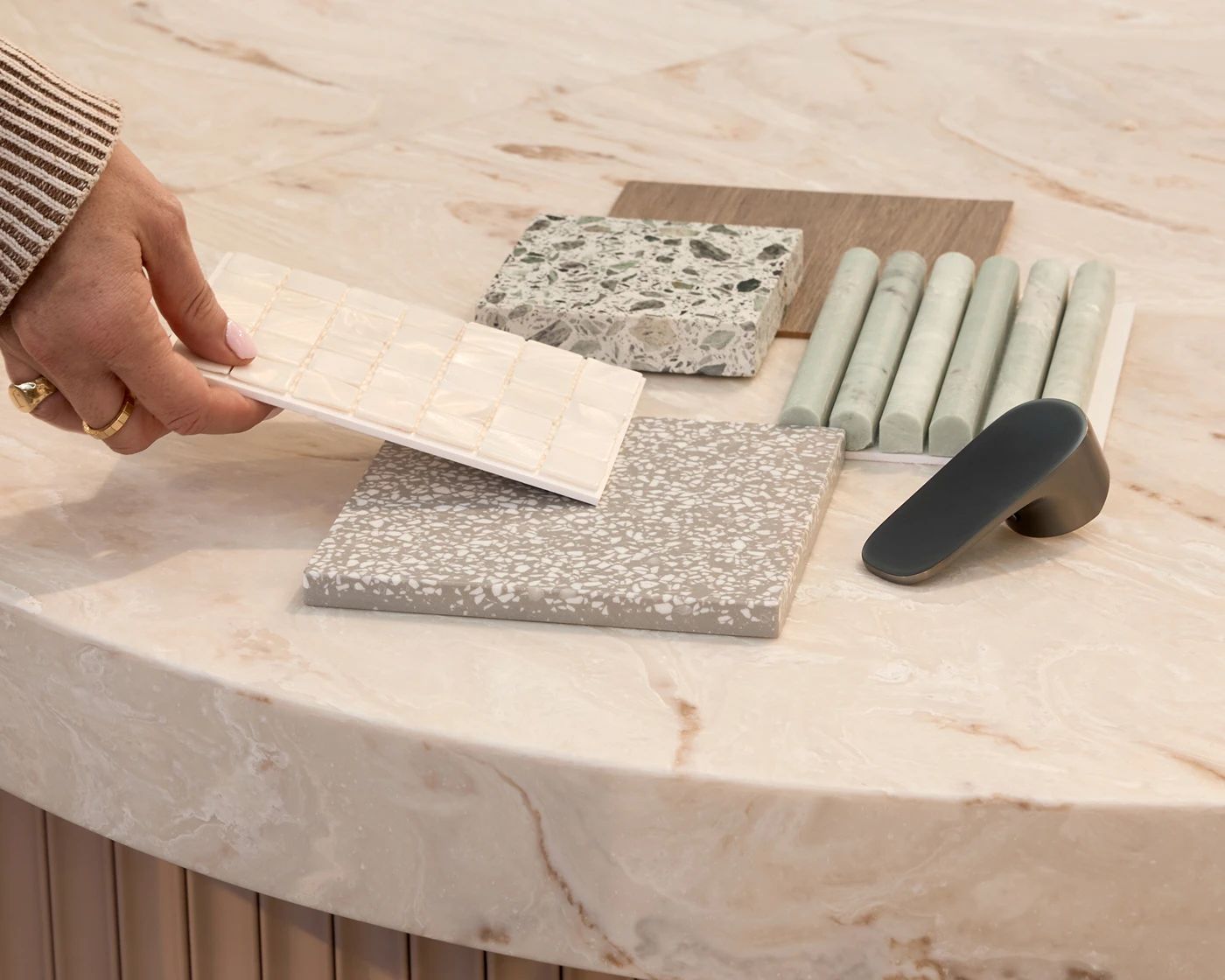 A hand holding a white tile sample over a marble surface with terrazzo, wood, and ceramic tile samples and a dark cabinet handle.