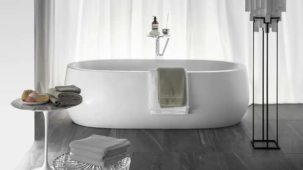 Modern white freestanding bathtub with towels and side tables in a minimalist bathroom with dark wood floors.