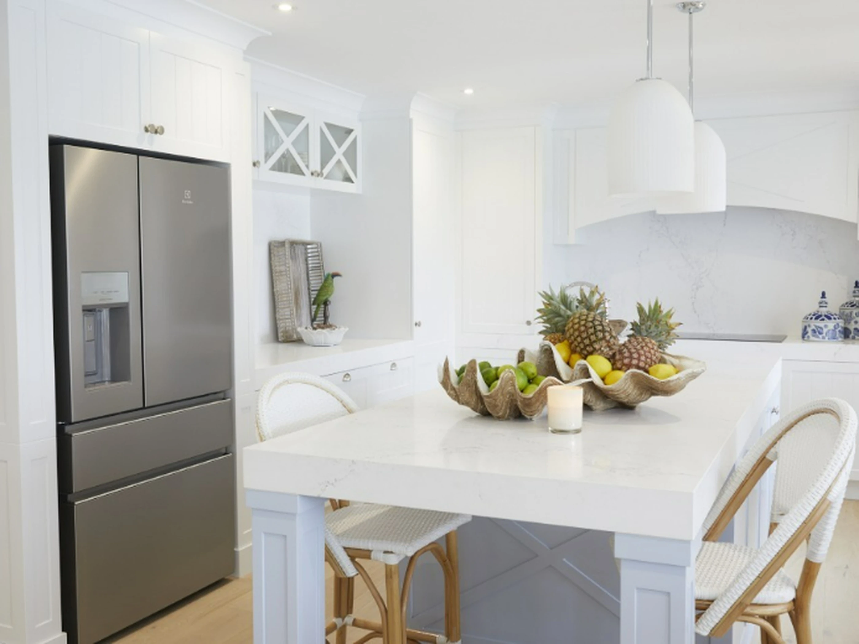 Modern white kitchen with stainless steel refrigerator, island featuring fruit bowl with pineapples, and white pendant lights.