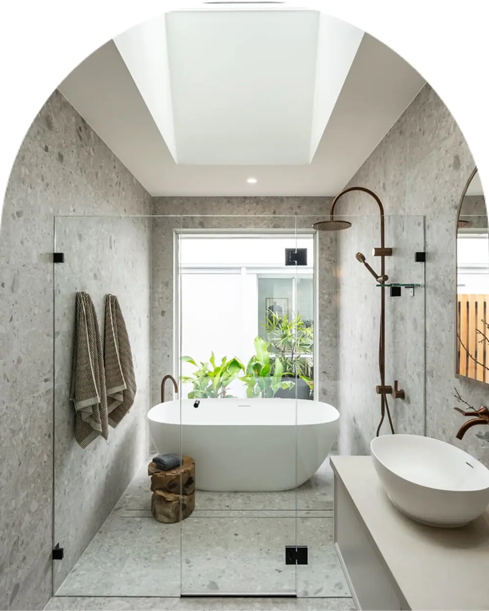 Modern bathroom with gray stone walls, white freestanding tub, skylight, and glass shower area overlooking green plants.