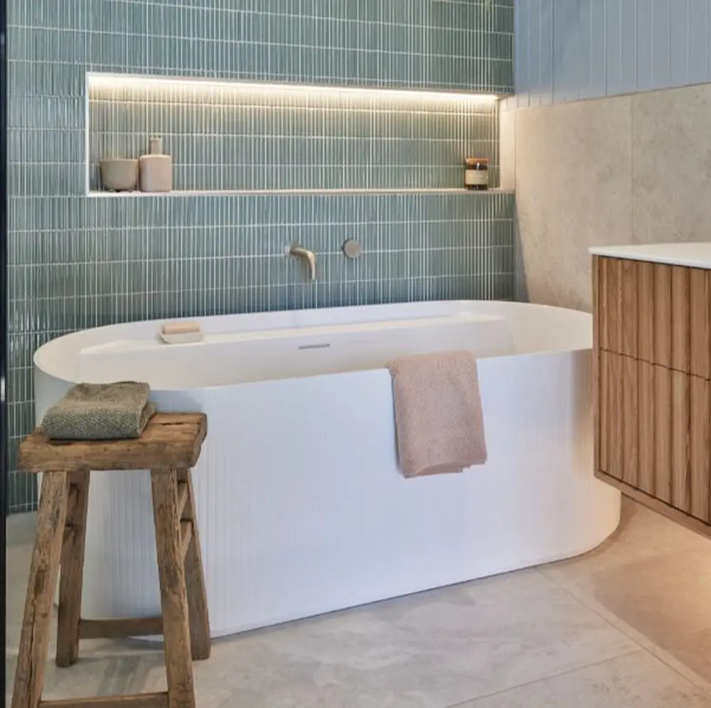 Modern bathroom with white freestanding tub, teal tile wall, wooden stool, and illuminated shelf niche.