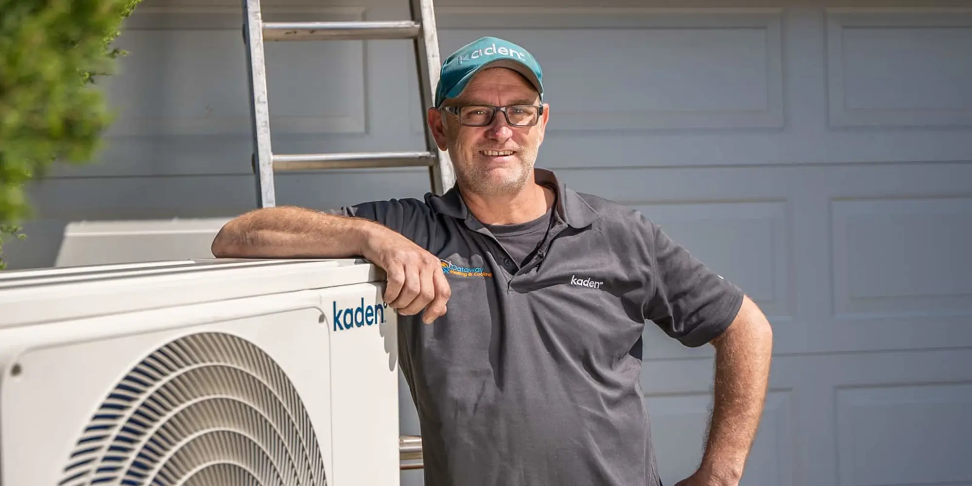 Person in gray Kaden shirt and teal cap standing beside air conditioning unit with ladder in background.
