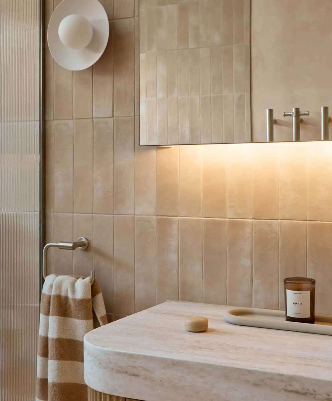 Modern bathroom with beige tiled walls, marble sink, striped towel, candle, and round wall light fixture.