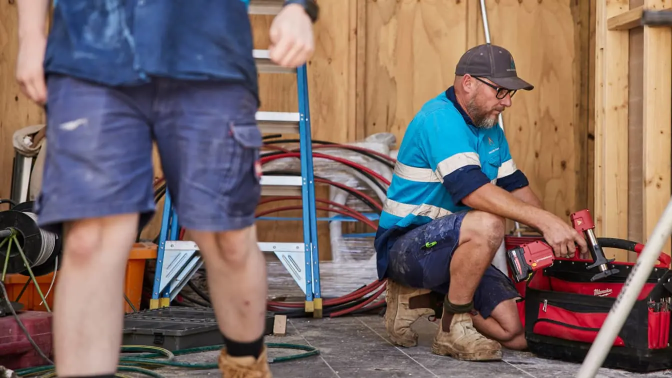 Construction workers in blue uniforms at a wooden-walled job site, one kneeling with tools and a red toolbag.