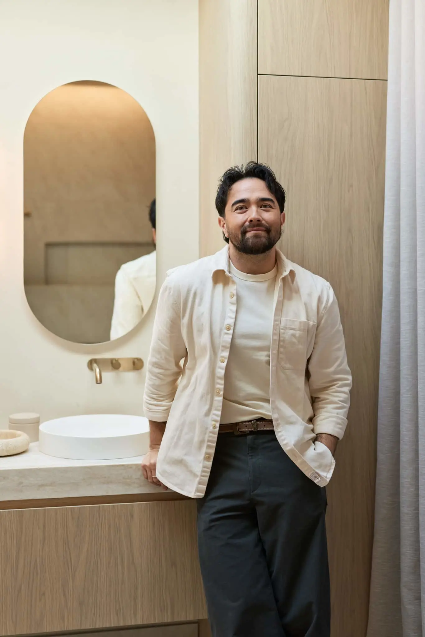 A man in a cream shirt and dark trousers leaning against a bathroom vanity with an oval mirror behind him.