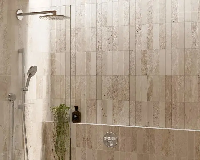Modern shower with beige stone-look tiles, rainfall showerhead, handheld sprayer, and small plant on built-in shelf.