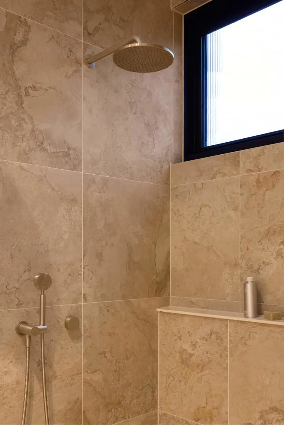 Beige tiled shower with brass rainfall showerhead, handheld sprayer, and built-in shelf with bottle near window.
