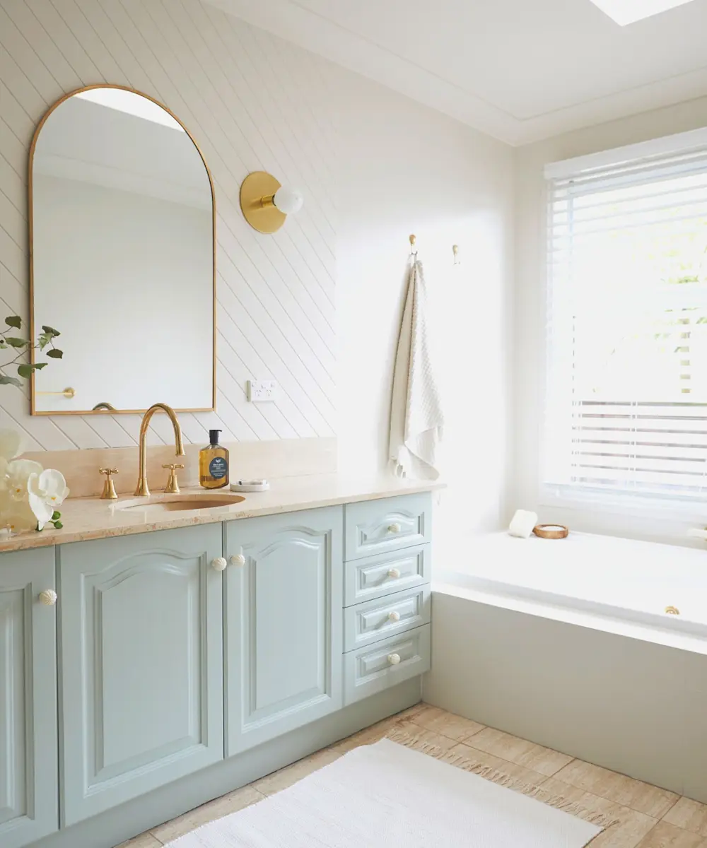Bright bathroom with mint green vanity, gold-framed arched mirror, brass fixtures, white bathtub, and natural light from window.