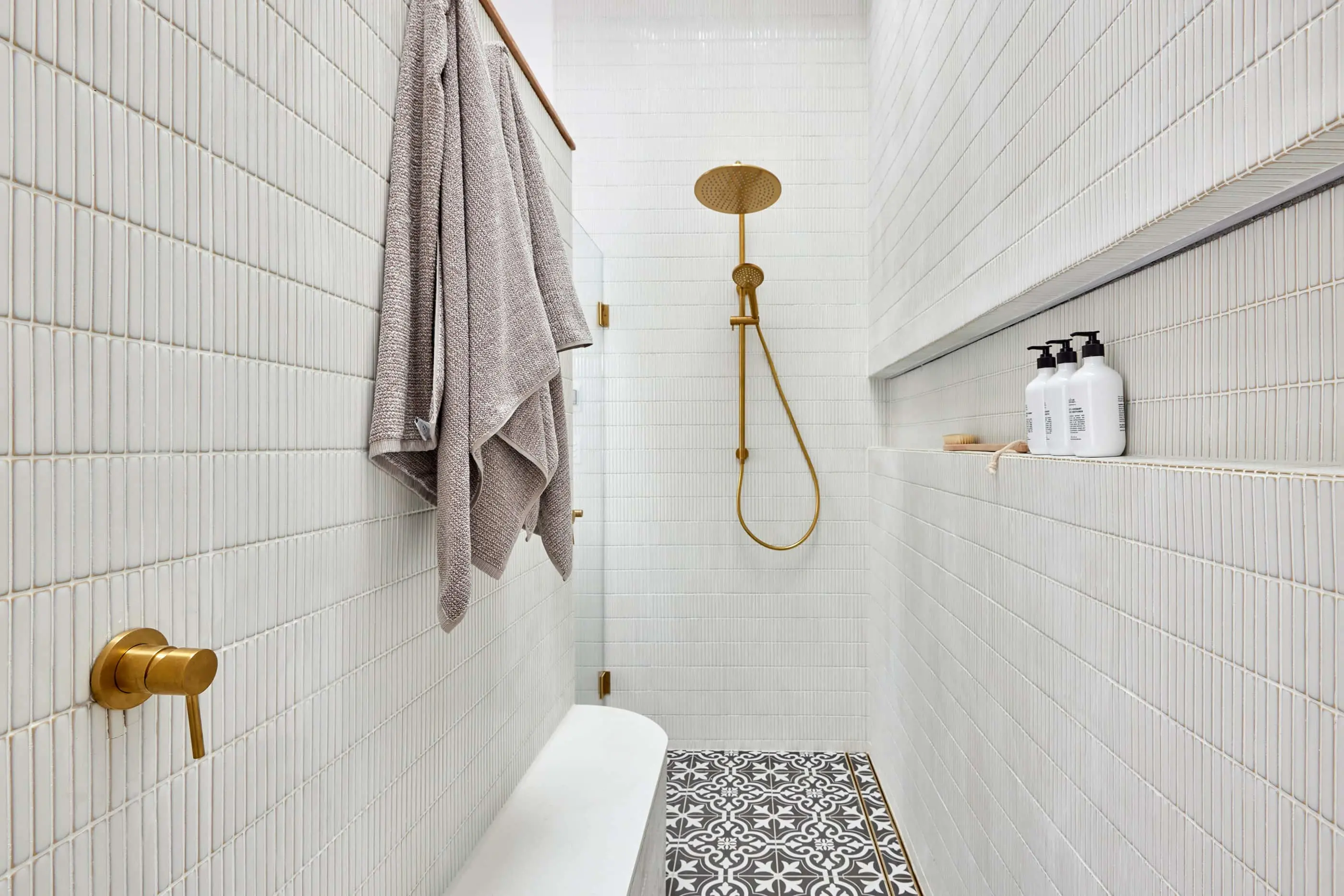 Modern white tiled bathroom with gold fixtures, hanging gray towel, patterned floor, and built-in shelf with toiletries.