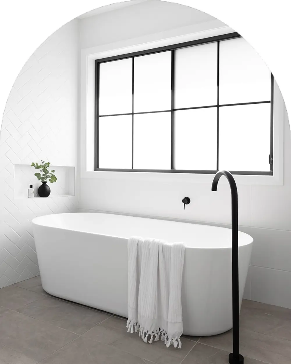 Modern white freestanding bathtub with black fixtures in a minimalist bathroom featuring a large window and small plant.
