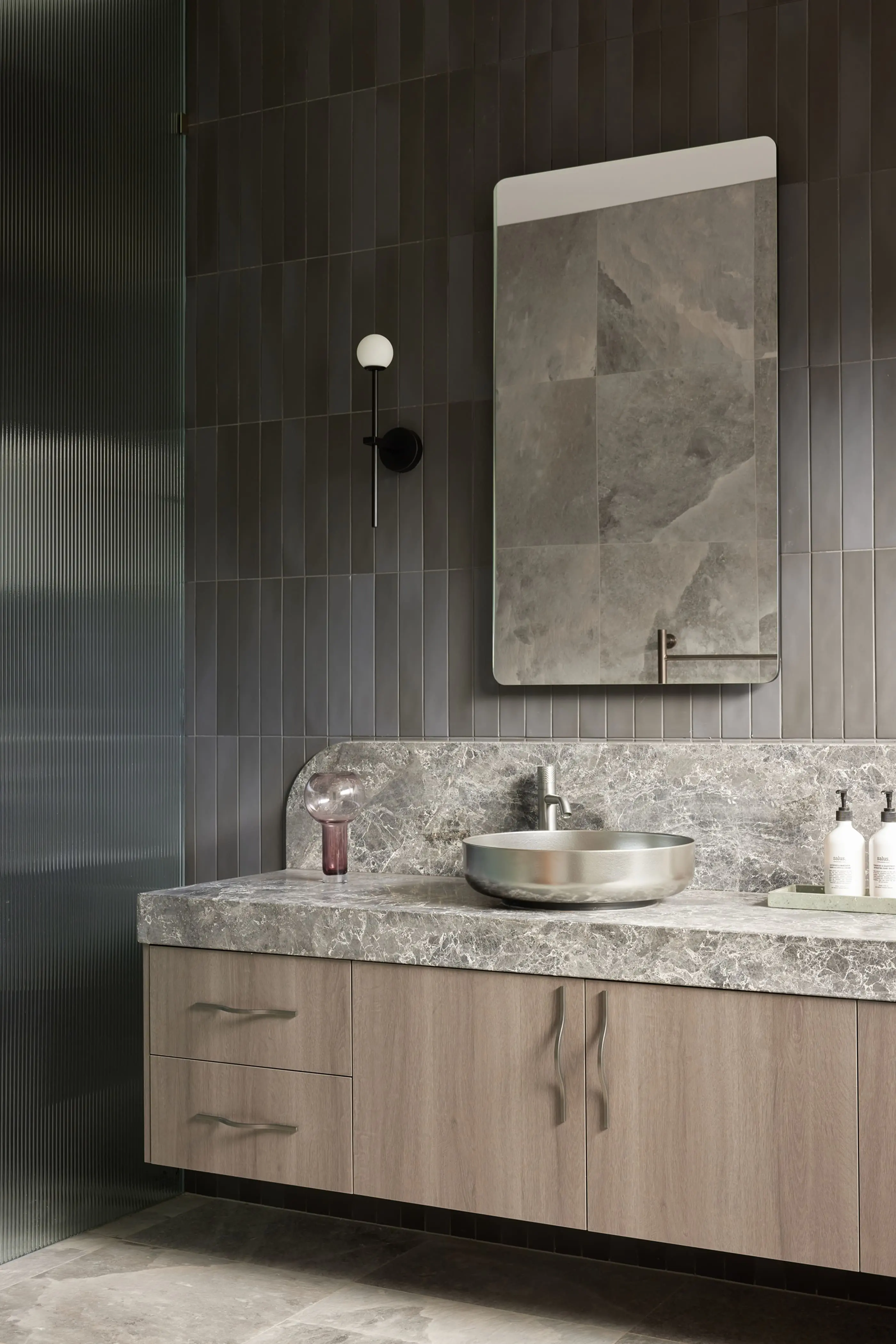 Modern bathroom with dark gray tile walls, floating wood vanity, marble countertop, metal vessel sink, and wall sconce.