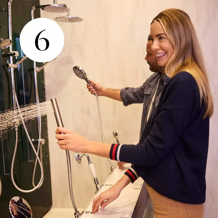 Two people testing handheld showerheads on display in a showroom, with water spraying from one showerhead.