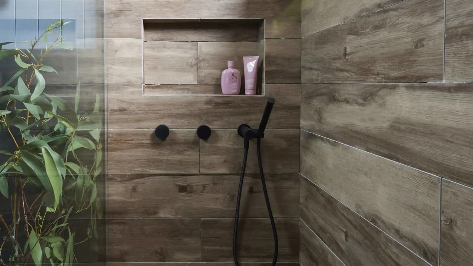 Modern shower with wood-look tiles, black fixtures, built-in shelf with pink toiletries, and green plant visible.