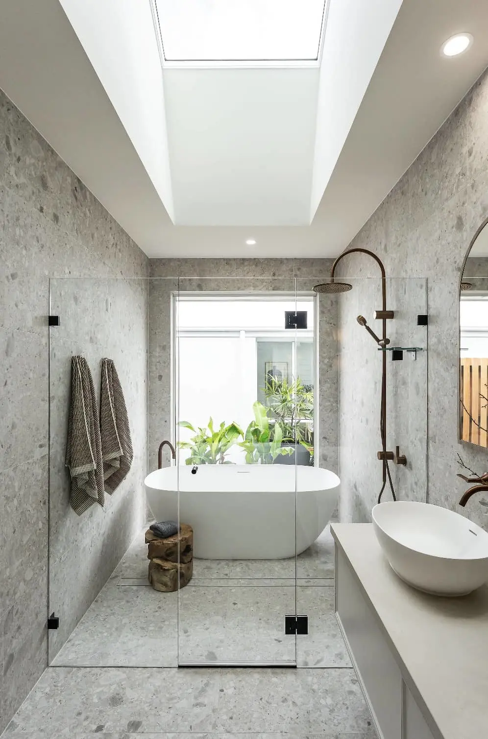 Modern bathroom with gray stone walls, white freestanding tub, skylight, and glass shower enclosure with plants visible outside.