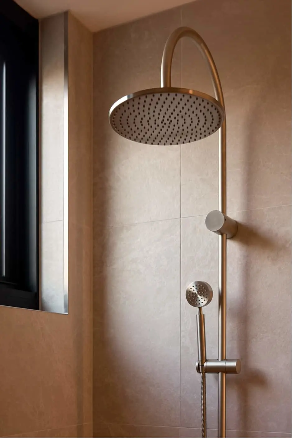 Brushed gold shower system with round rainfall head and handheld sprayer mounted on beige tiled wall.