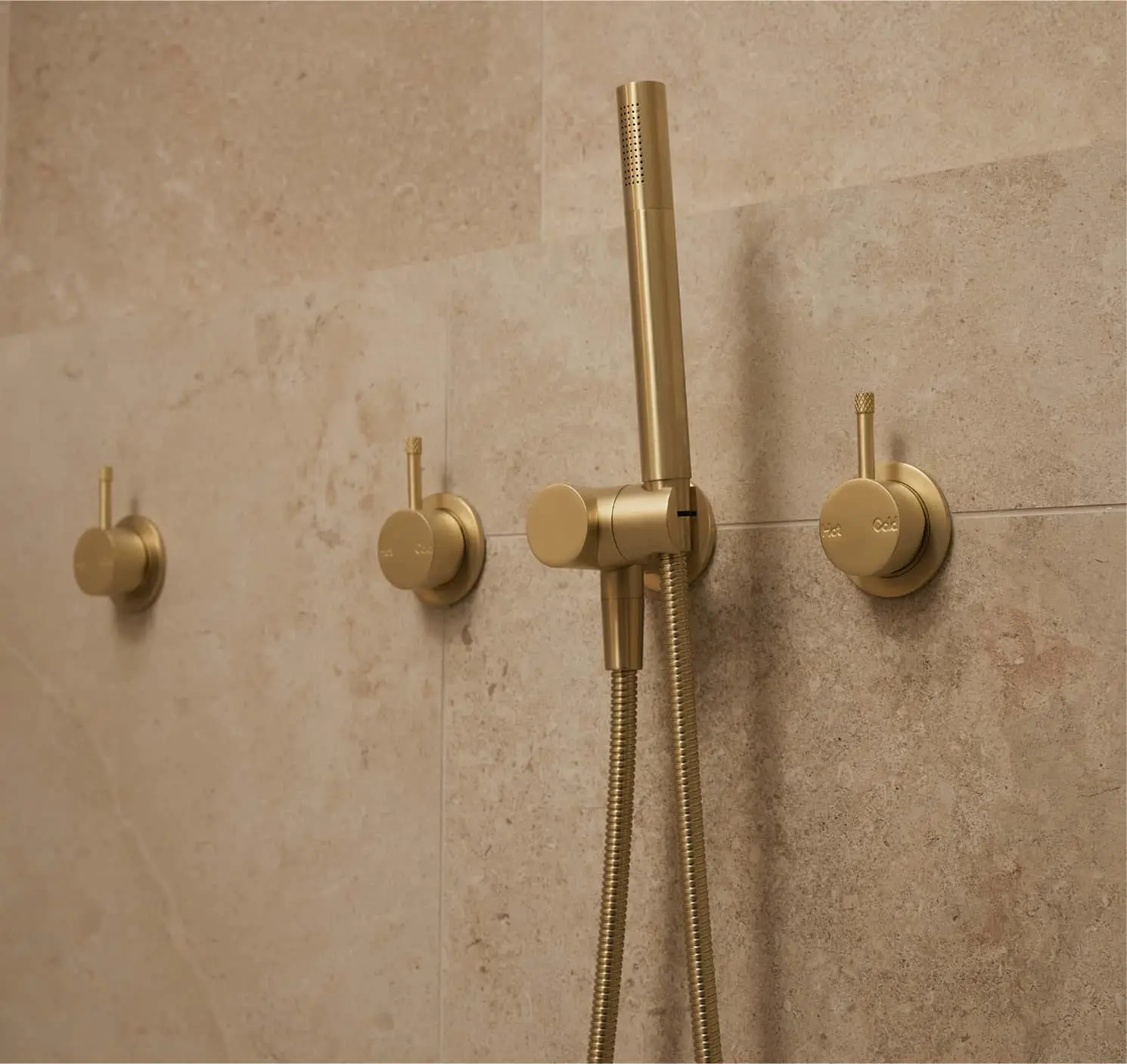 Brushed gold shower fixtures with handheld showerhead and hose mounted on beige tiled wall.