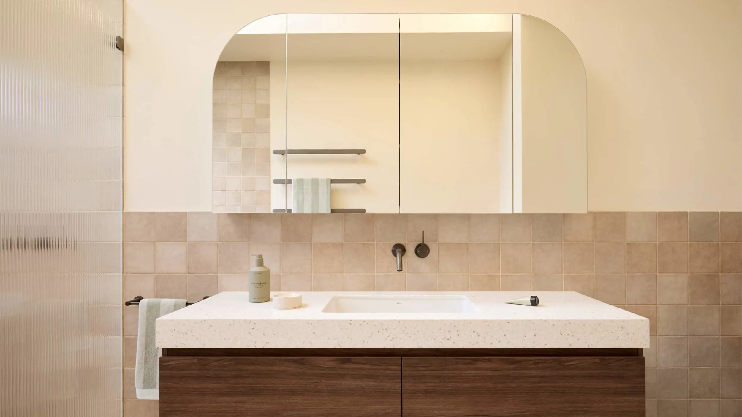 Modern bathroom with white sink, wooden vanity, beige tiled walls, and arched mirror cabinet above.