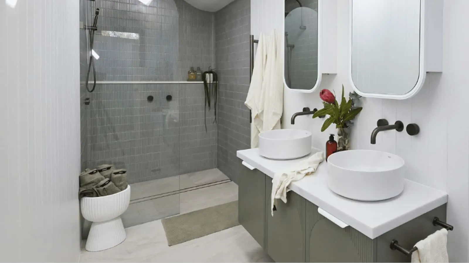Modern bathroom with gray tile shower, double white vessel sinks, round mirrors, and a small red flower in a vase.