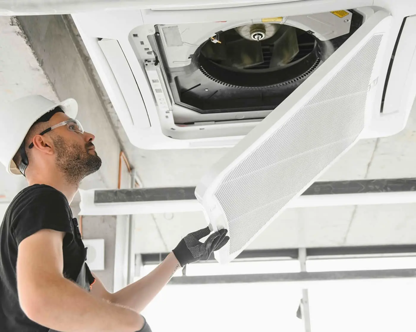 Technician in hard hat installing or inspecting ceiling air conditioning unit with exposed fan component.