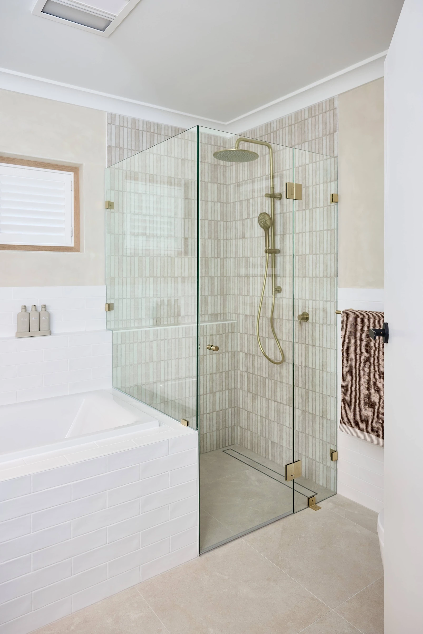 Modern bathroom with glass shower enclosure featuring brass fixtures, white bathtub, and beige tiled walls.