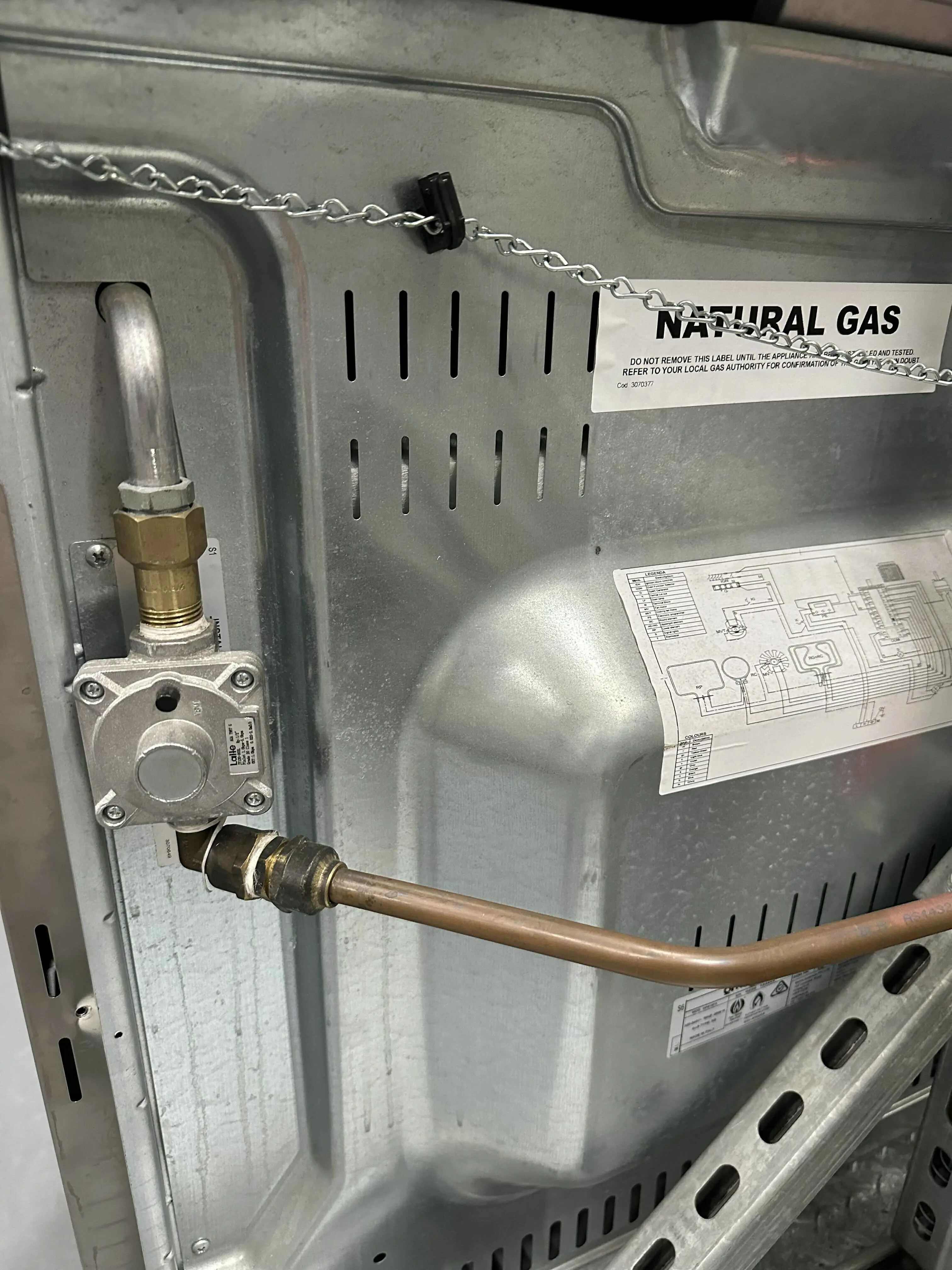 Natural gas connection on metal appliance showing valve, copper pipe, and warning label with wiring diagram.