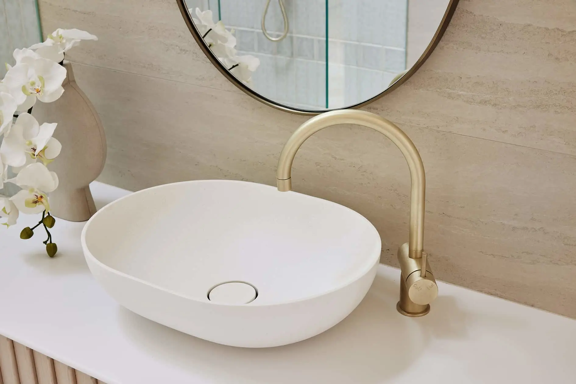 Modern bathroom with white vessel sink, gold faucet, round mirror, and white orchid on beige countertop.