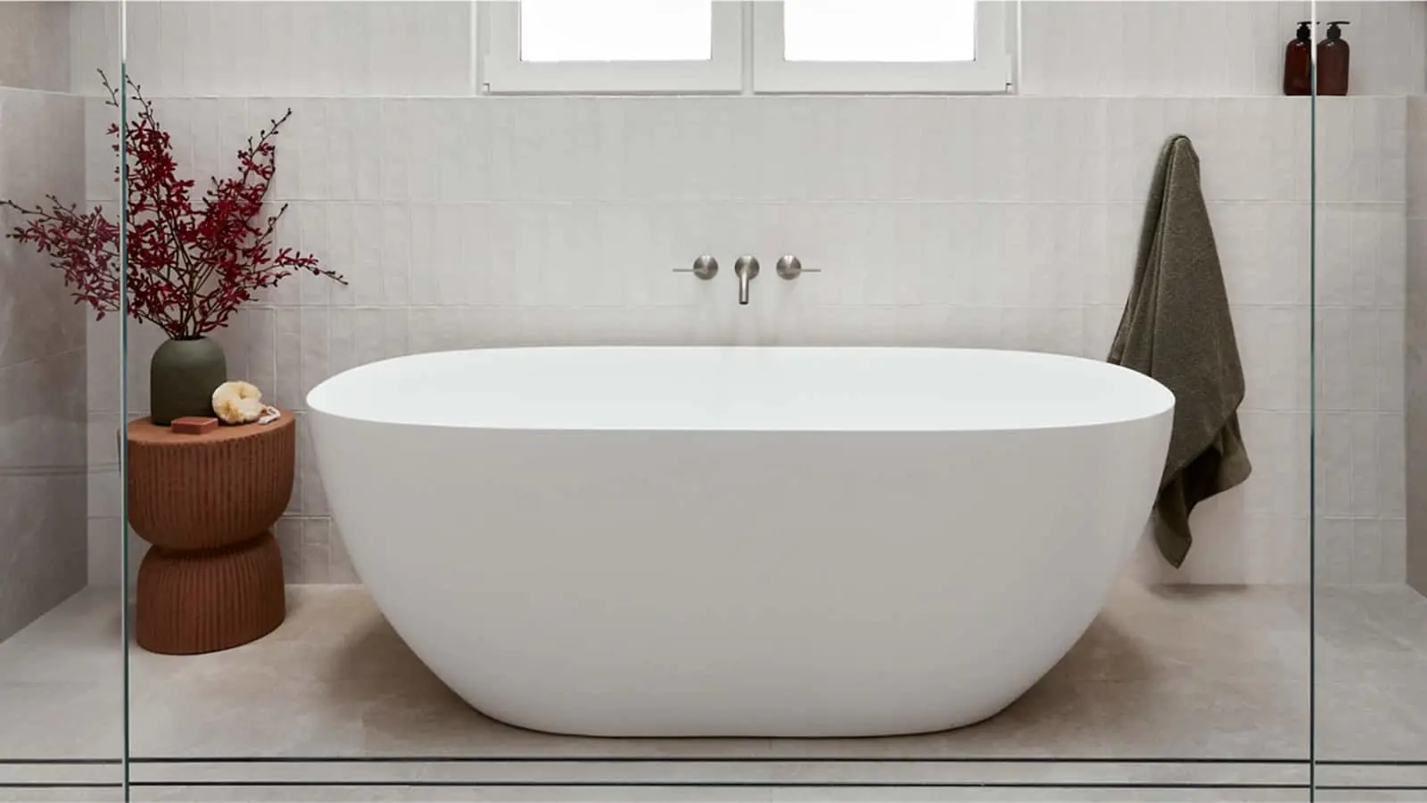 Modern white freestanding bathtub in a minimalist bathroom with red branches in a vase and wooden side table.