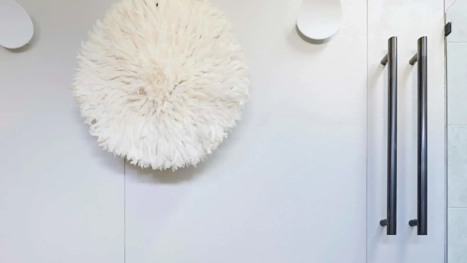 Circular white fluffy wall decoration next to modern cabinet with metal handles on a minimalist white wall.