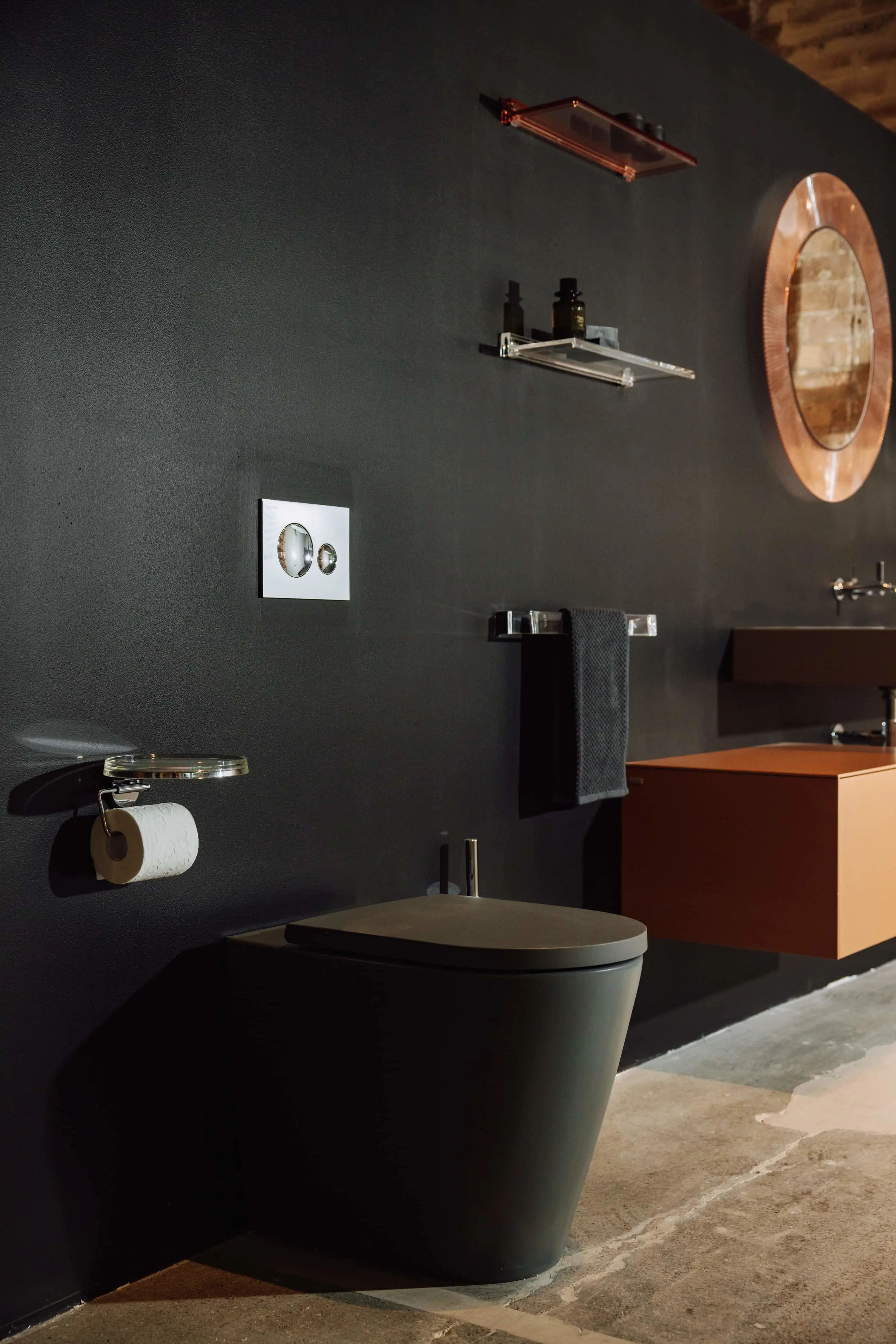 Modern bathroom with black walls, gray toilet, copper round mirror, glass shelves, and wooden vanity cabinet.