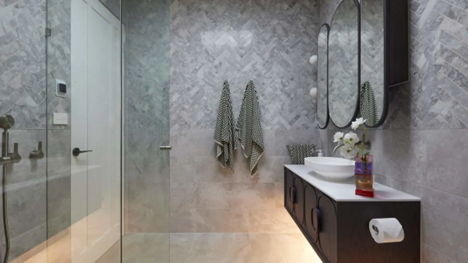 Modern bathroom with marble herringbone tile, glass shower, black vanity with white sink, oval mirror, and patterned towels.