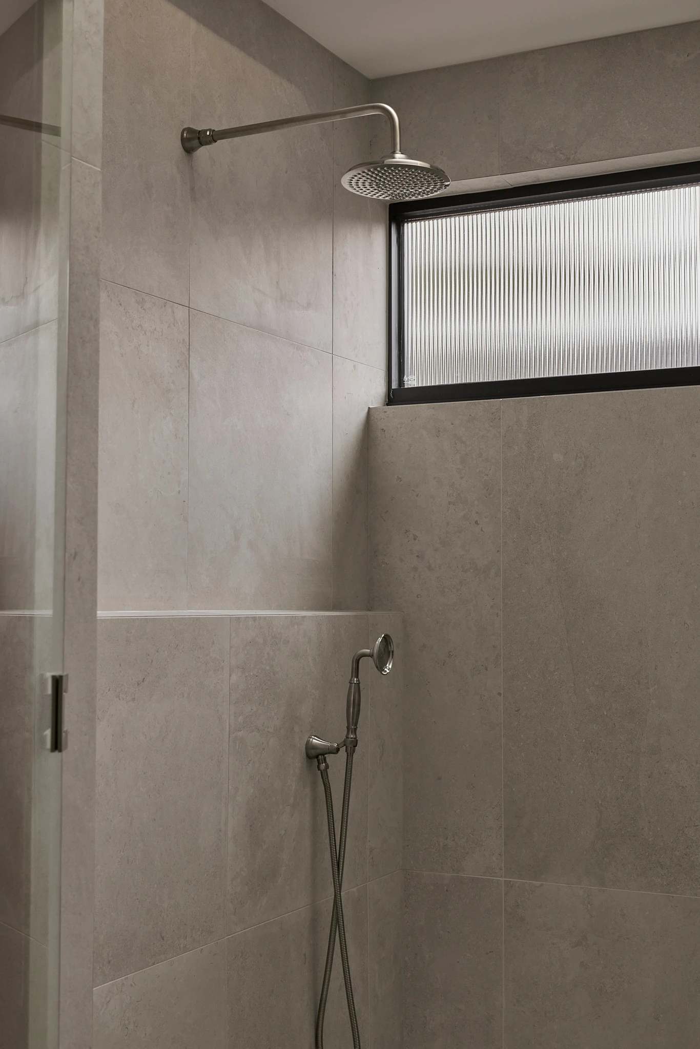 Modern shower with beige tile walls, rainfall showerhead, handheld sprayer, and ribbed glass window for privacy.