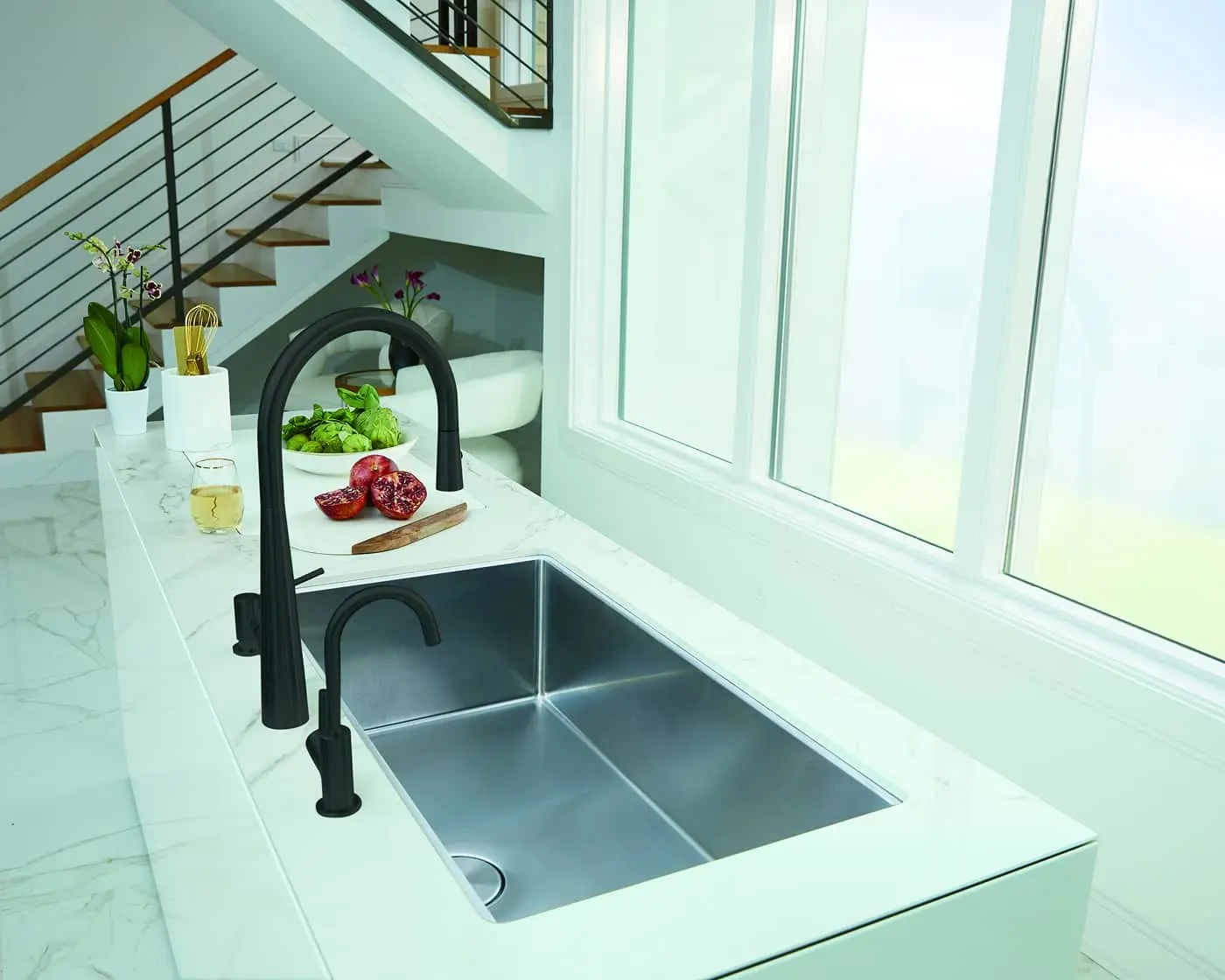 Modern kitchen with black faucet, stainless steel sink, marble countertop, fresh produce, and staircase in background.