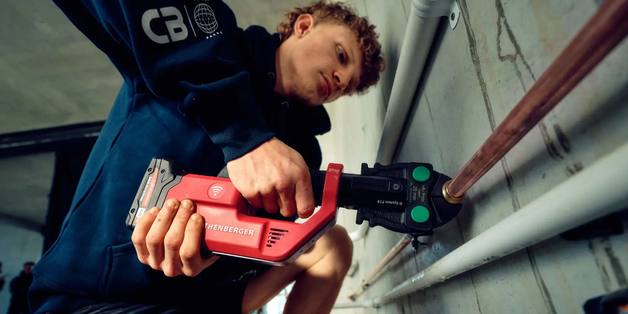 Person in blue jacket using red Rothenberger crimping tool on copper pipe with green button attachment.