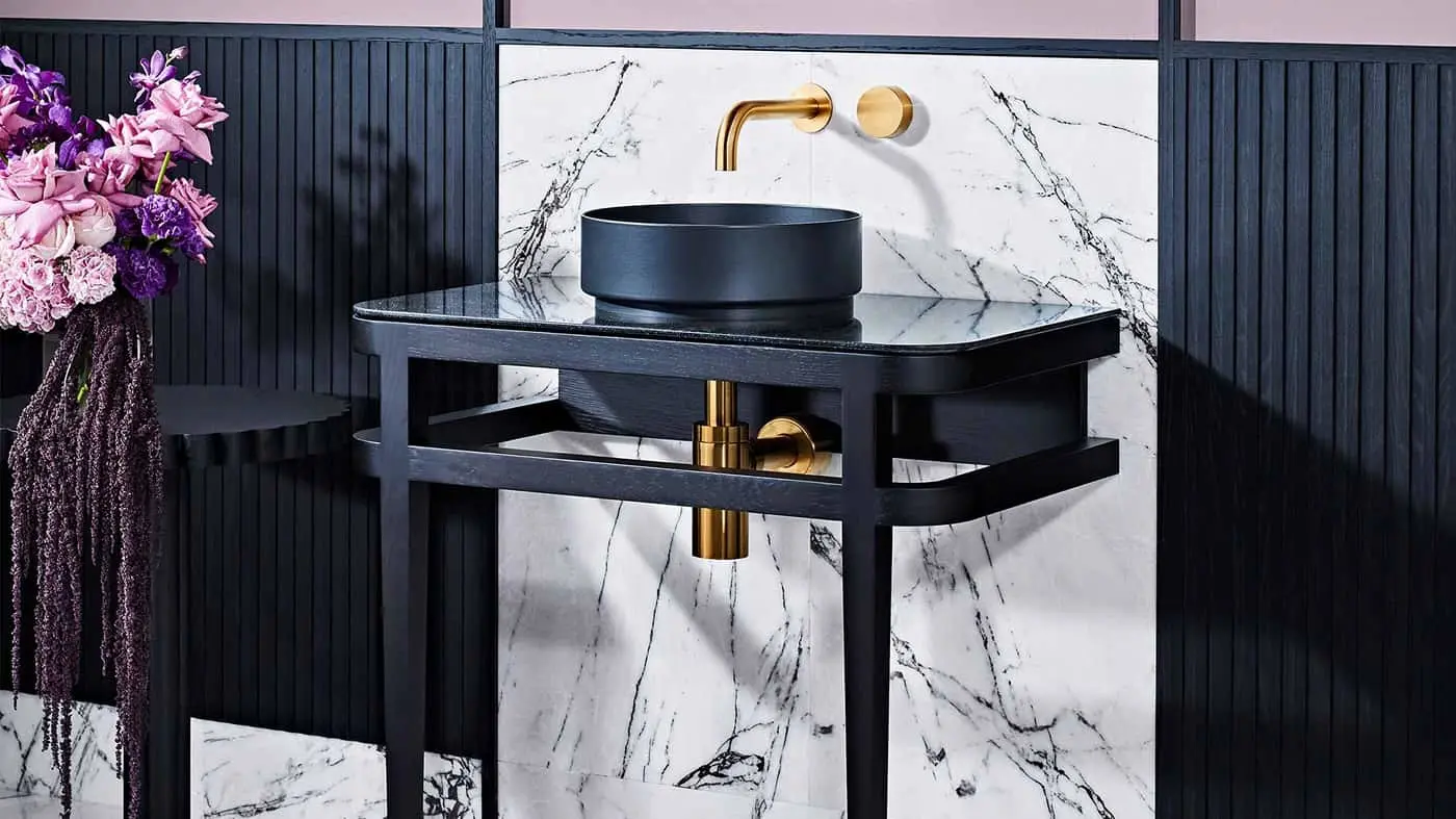 Modern bathroom vanity with black basin on dark wood stand, brass fixtures, marble wall, and pink-purple flowers nearby.