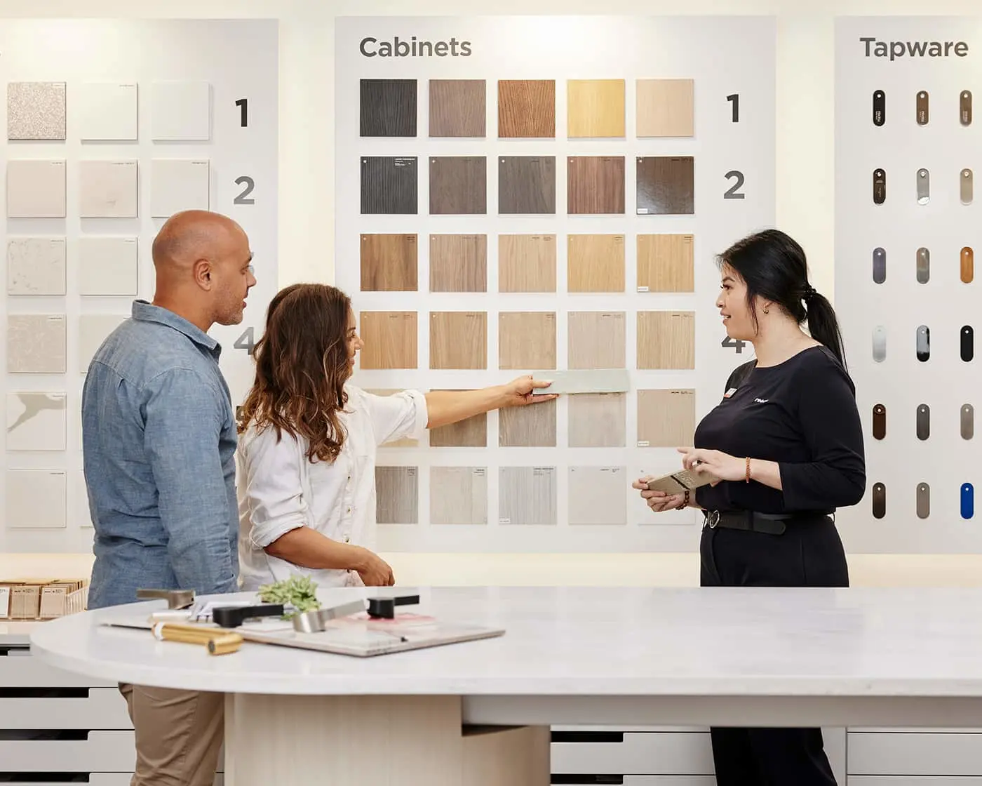 Couple selecting cabinet finishes with design consultant in showroom displaying wood samples and hardware options.