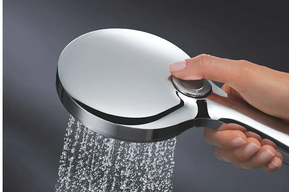 Hand holding a modern chrome showerhead with water spraying from its flat, circular design against a dark background.