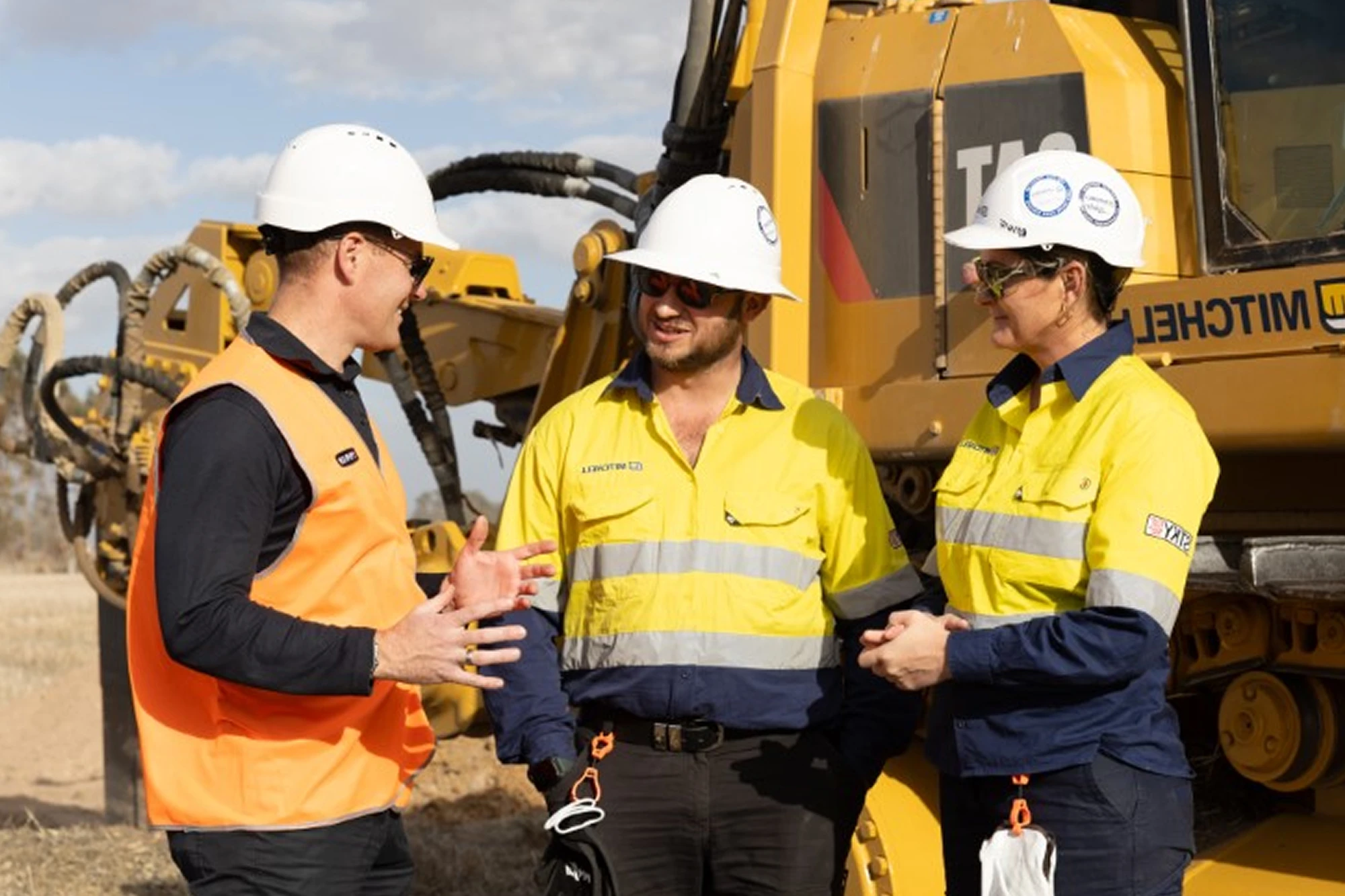 Three construction workers in hard hats and high-visibility clothing discussing work near a yellow excavator.