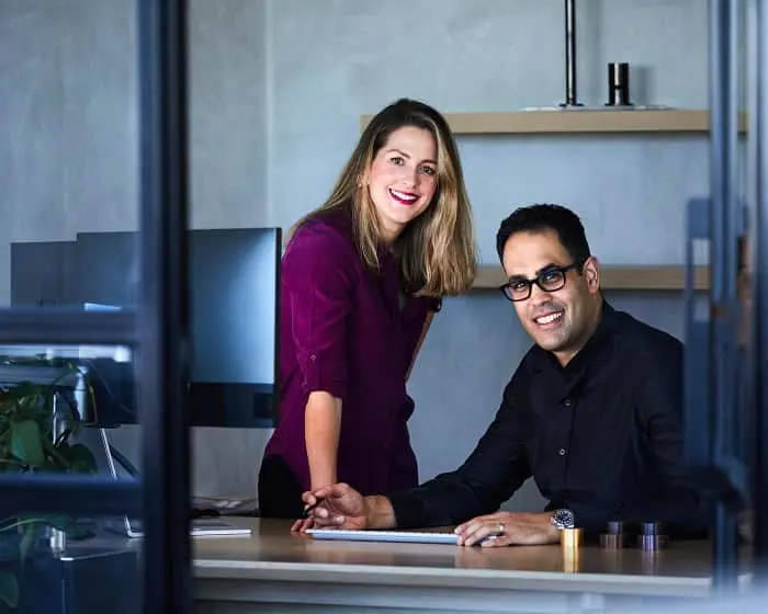 Two smiling professionals in a modern office, woman in purple top and man in black shirt sitting at desk.