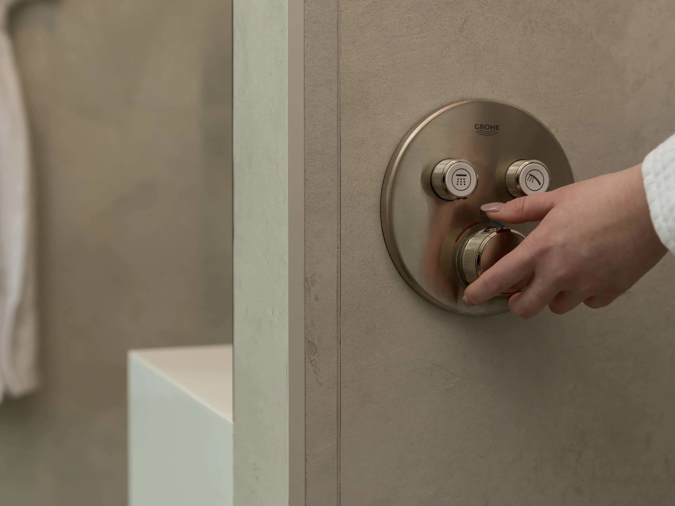 Hand adjusting a brushed metal Grohe shower control panel with multiple dials mounted on a beige bathroom wall.