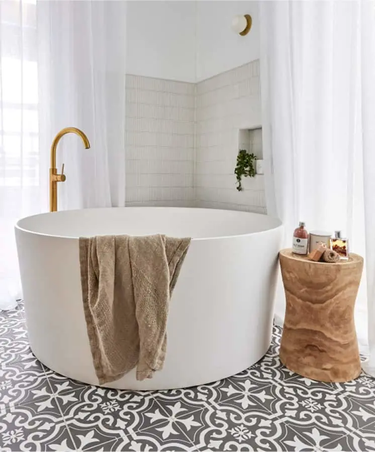 Modern white circular bathtub with brass faucet, beige towel, wooden side table, and patterned floor tiles in a bright bathroom.