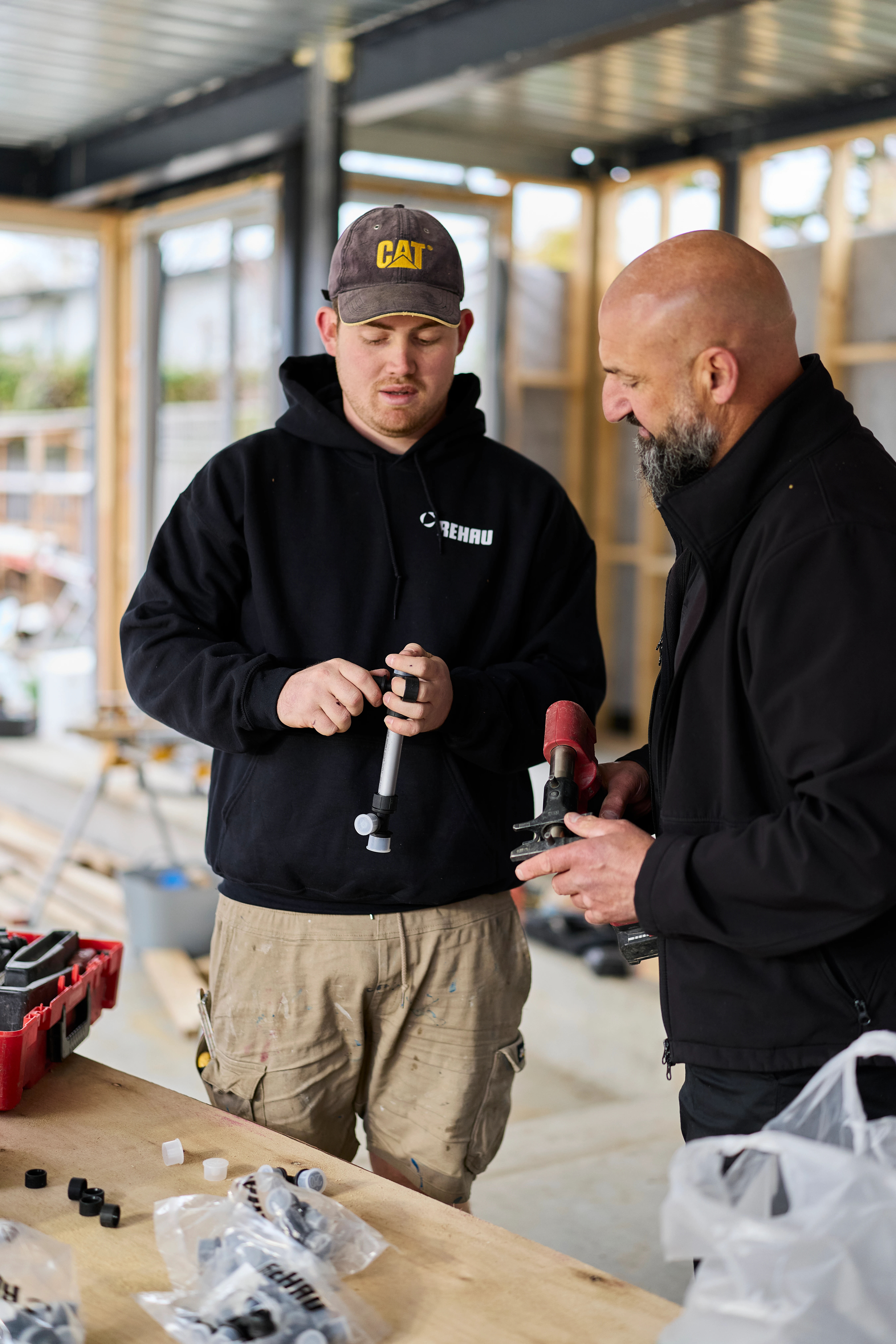 Two construction workers examining tools at a worksite, one wearing a CAT cap and black hoodie, standing at a wooden table.