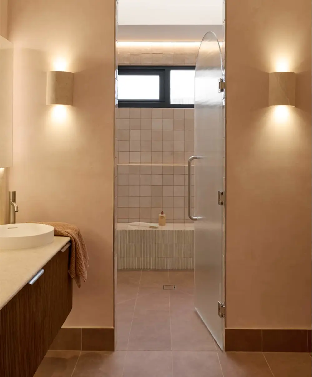 Modern bathroom with warm beige walls, vessel sink, and glass shower door. Soft lighting and minimalist design.