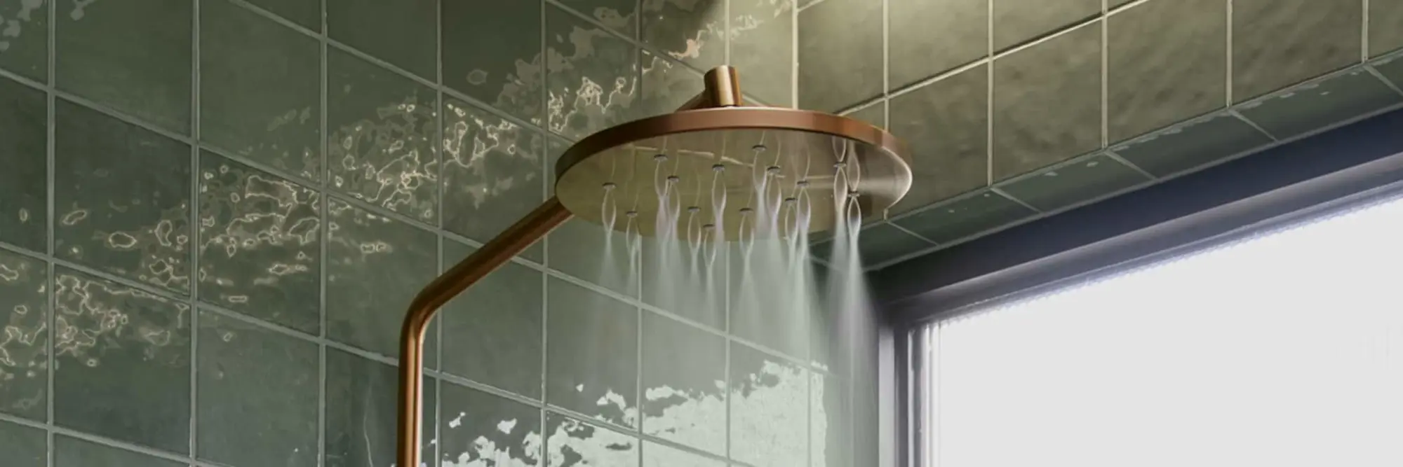 Copper rainfall showerhead with water flowing, mounted on green tiled bathroom wall next to a window.