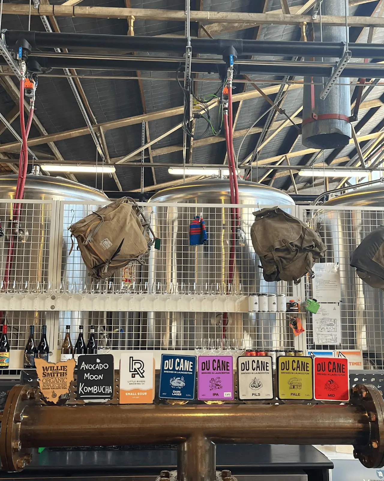 Brewery interior with beer taps featuring Du Cane and other craft beers, vintage bags hanging above stainless tanks.