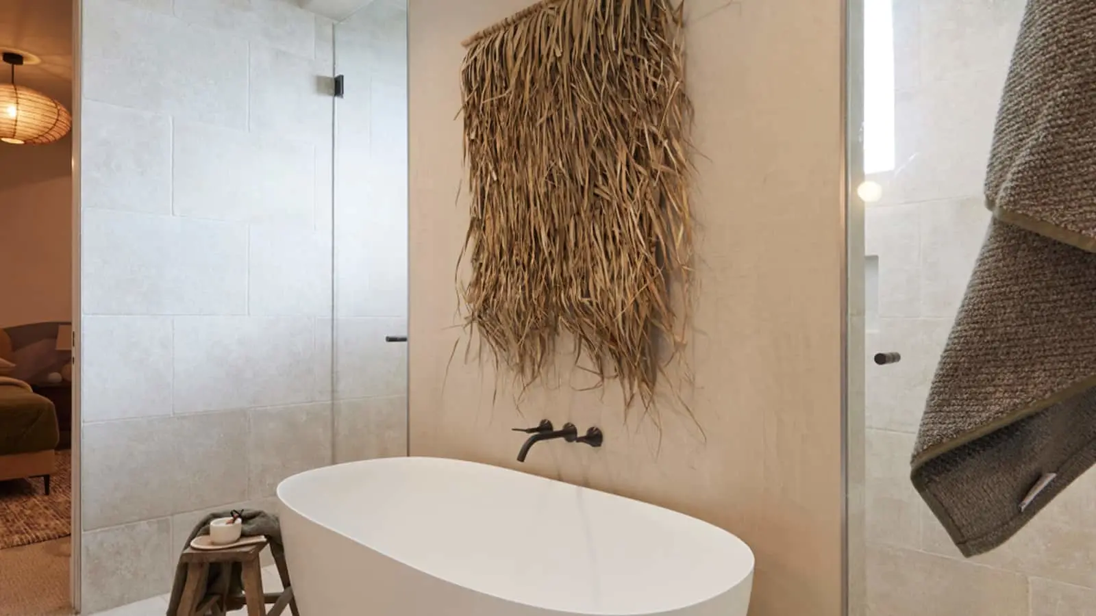 Modern bathroom with white freestanding tub, natural fiber wall hanging, and glass shower door.