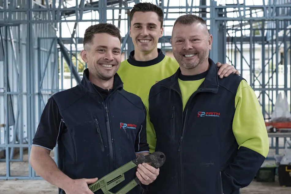 Three construction workers in navy vests and hi-vis shirts smiling at a building site with metal framing in background.